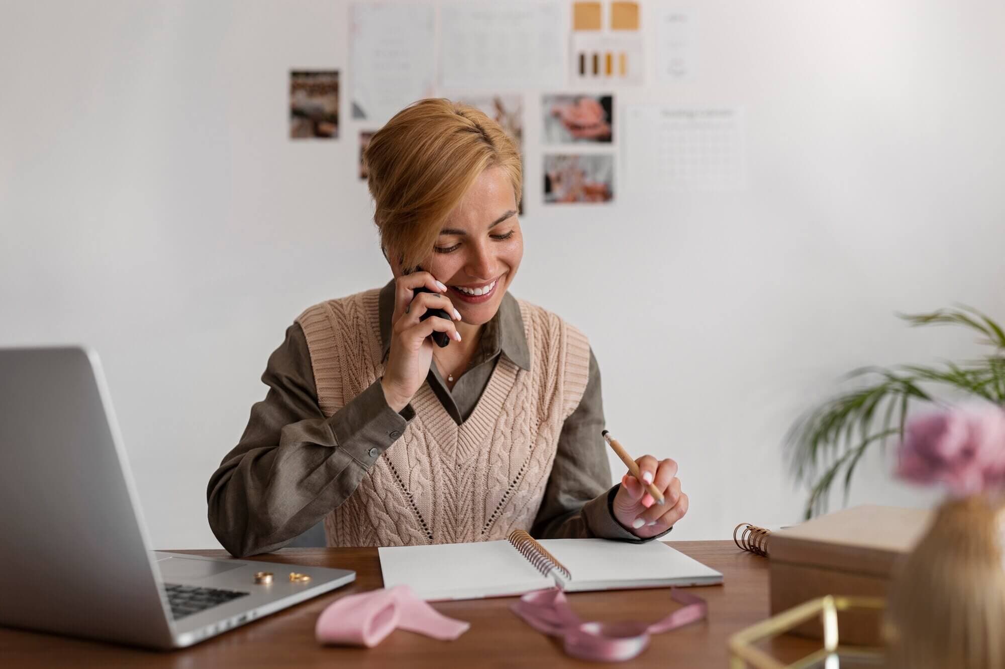 A woman works at a laptop while talking on her phone and reviewing plans.