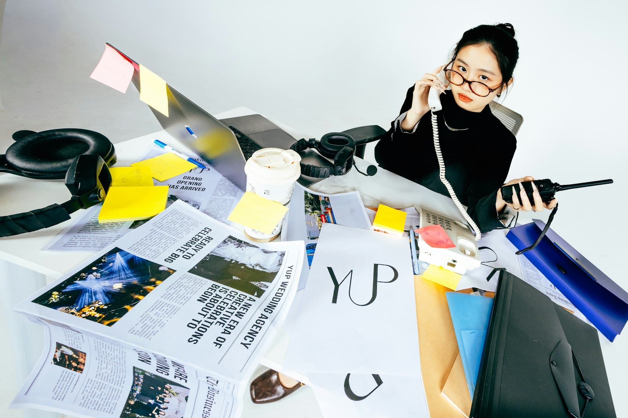 A woman sits at a desk with papers, a laptop, and a phone in hand.